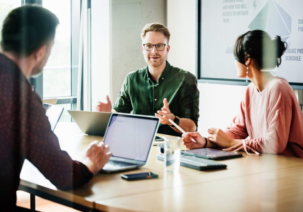 A focused team leader is addressing colleagues in a meeting, with a presentation displayed in the background, capturing a moment of active corporate engagement.