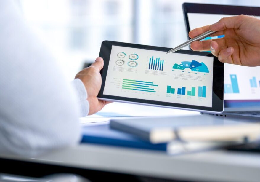 Two business men meeting and looking at financial data, charts and graphs on a digital tablet and a laptop computer. There are documents and paperwork on the table. One man is pointing at the screen